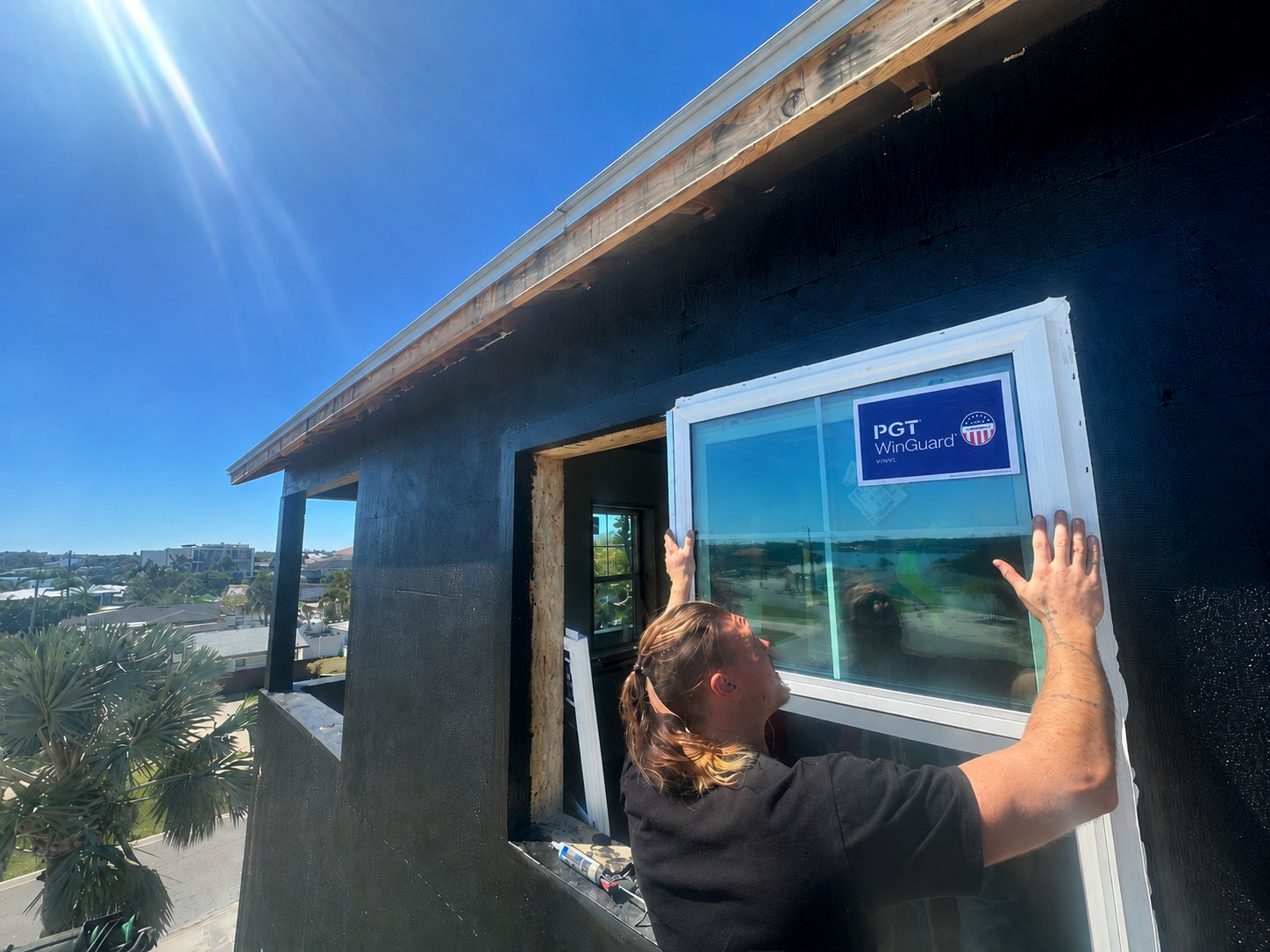 A Hausmark installer setting a PGT WinGuard vinyl impact window into a prepared opening, the water behind the house